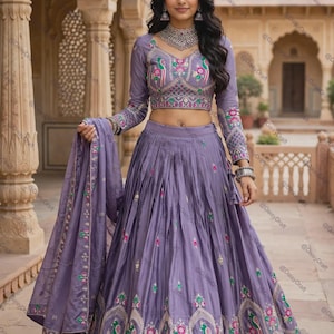May include: A lavender lehenga choli set featuring a long-sleeved blouse and a pleated floor-length skirt. The garment is adorned with intricate embroidery in pink, green, and white, showcasing peacock motifs and geometric patterns along the hem.