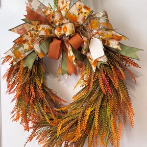 May include: A decorative wreath featuring a base of dense, feathery foliage in shades of orange, yellow, and green. The top is adorned with a large, multi-layered bow made of patterned ribbon featuring pumpkins and a solid orange glittered ribbon.
