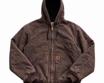 May include: A dark brown zip-up hooded jacket with a black interior lining. The jacket features a front zipper closure, two front pockets, ribbed cuffs and hem, and a small rectangular Carhartt brand patch on the lower front pocket area.
