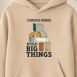 May include: A beige hooded sweatshirt featuring a graphic print of stacked wooden building blocks in various shapes and colours. The text CURIOUS MINDS BUILD BIG THINGS is printed in dark blue lettering above and below the illustration of the blocks.