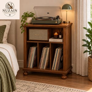 May include: A wooden record player stand with a rectangular top, four bun feet, and three vertical storage compartments for vinyl records. The top surface holds a green turntable, while the middle shelf contains a Marshall speaker and a book.