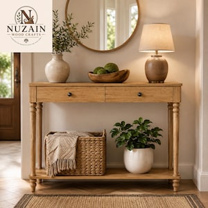 May include: A light brown wooden console table with two drawers, four turned legs, and a lower shelf. On top sits a beige vase, a wooden bowl with green spheres, and a lamp. Below, a woven basket with a beige throw and a white potted plant.