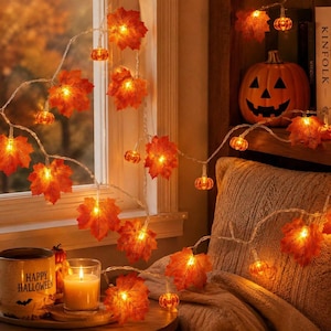 May include: A string of LED fairy lights featuring orange maple leaves and small pumpkin-shaped bulbs. The lights emit a warm glow, illuminating the artificial foliage and miniature pumpkins. A candle in a mug labelled Happy Halloween is nearby.