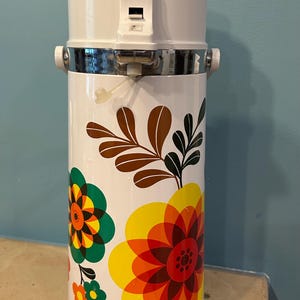 May include: A white cylindrical thermal beverage dispenser featuring a colorful floral pattern with overlapping petals in shades of yellow, orange, red, and green, along with brown leaf motifs. The top includes a white pump mechanism and handle.