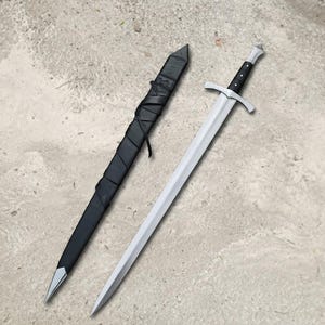 May include: A silver-colored metal sword with a black handle and a silver pommel, positioned next to a black scabbard. The scabbard features a pointed silver tip and is wrapped in black leather straps that are tied around the exterior surface.