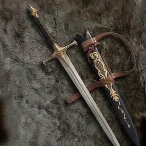 May include: A silver-toned sword with a gold-colored crossguard and pommel, featuring a dark brown wrapped grip. Beside it lies a black scabbard adorned with gold-colored filigree patterns and attached brown leather straps for carrying.