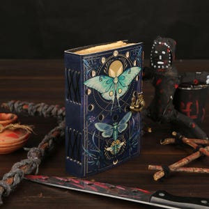 Peut inclure: Un journal relié en cuir bleu foncé orné d'une illustration de papillon de nuit, de graphismes de phases lunaires et d'un fermoir en métal doré. Le dos est cousu avec un cordon noir et la couverture présente des motifs célestes complexes.