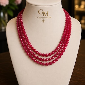 Op de afbeelding: Een ketting met drie strengen bestaande uit gepolijste, ronde, dieproze kralen. De ketting wordt getoond op een beige buste met de gouden tekst GM Les Pierres de GM en een klein ruitvormig icoon boven de ketting.