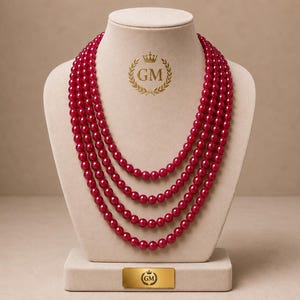 May include: A multi-strand necklace featuring four layers of polished, deep red spherical beads. The necklace is displayed on a beige bust. A gold-coloured logo with the letters GM and a crown icon is visible on the bust and on a rectangular plaque.