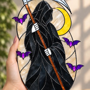 May include: An oval stained glass panel featuring a hooded figure in a black robe holding a scythe with a silver blade and orange handle. The background includes a yellow crescent moon and four small purple bats, all outlined in black metal cames.