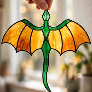 May include: A stained glass suncatcher shaped like a dragon with a green body and translucent orange wings. The piece features dark metal soldering lines outlining the wings and body, creating a segmented appearance with vibrant colors.