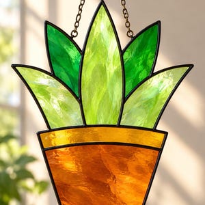 May include: A stained glass suncatcher shaped like a potted plant with vibrant green leaves and an amber-colored pot. The piece features black metal framing and is suspended by a thin metal chain for hanging in a window or light source.