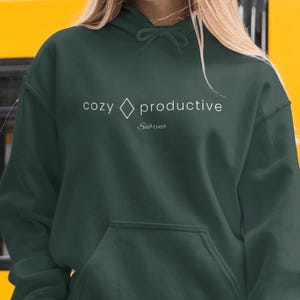 Könnte beinhalten: Ein dunkelwaldgrüner Kapuzenpullover mit einer Kängurutasche auf der Vorderseite und passenden Kordelzügen. Auf der Brust befindet sich der weiße Schriftzug "cozy", gefolgt von einer Raute, dem Wort "productive" und dem Namen "Sabrina".