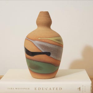 May include: A ceramic vase with a narrow, ribbed neck and a rounded body. The surface features a matte, tan base color decorated with abstract, flowing bands of sage green, slate gray, and glossy black glaze, creating a layered, organic appearance.