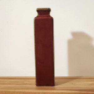 May include: A tall, rectangular ceramic vase with a deep matte red finish and contrasting gold-toned edges along the vertical sides and the rim. The vase features a narrow neck and a flat, square-shaped opening at the top, standing upright.