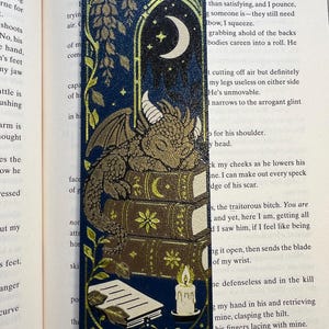 Könnte beinhalten: Ein rechteckiges Lesezeichen mit dunkelblauem Hintergrund und Goldfolien-Illustrationen. Das Design zeigt einen kleinen Drachen, der auf einem Stapel von drei Büchern schläft, eine Mondsichel in einem Bogenfenster, Ranken und eine brennende Kerze.