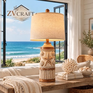 Puede incluir: Una lámpara de mesa con pantalla cilíndrica de lino beige y base con relieve de estrella de mar, conchas y detalles de cuerda. La base incluye una sección inferior con listones de madera verticales y un acabado de tono cálido.