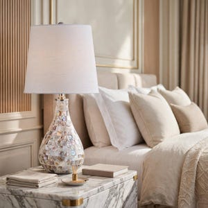 May include: A table lamp with a white fabric tapered shade and a pear-shaped base covered in iridescent, multi-colored mother-of-pearl shell tiles. The lamp features a metallic neck and sits on a white marble surface next to a small tray.