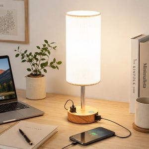 May include: A cylindrical table lamp with a white fabric shade and a light wood base. The base features a silver metal stem, a power button, and two USB ports. A black charging cable connects the lamp to a smartphone resting on the surface.