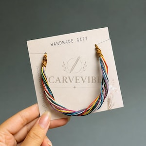 May include: A multi-colored string bracelet featuring thin, twisted strands in shades of red, yellow, green, blue, and white. The bracelet is attached to a square off-white display card with the text HANDMADE GIFT and CARVEVIBE printed on it.