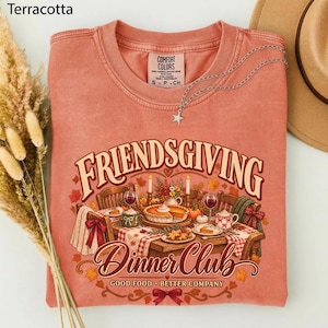 May include: A terracotta-colored crewneck t-shirt featuring a graphic of a dining table set for a meal. The text reads "FRIENDSGIVING DINNER CLUB" and "GOOD FOOD BETTER COMPANY". A silver star necklace rests on the collar of the shirt.