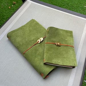 Op de afbeelding: Twee olijfgroene suède notitieboeken met oranje elastieken en ronde koperen bedels. Het grotere notitieboek heeft drie koperen bedels aan het elastiek, terwijl het kleinere notitieboek er twee heeft. Beide notitieboeken zijn afgewerkt met oranje stiksels.