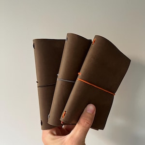 Puede incluir: Tres cuadernos de cuero marrón sostenidos juntos, cada uno con una banda elástica de cierre distinta en marrón oscuro, gris y naranja brillante. Las cubiertas tienen una textura mate suave y están aseguradas con cordones elásticos.