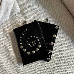 Puede incluir: Dos cuadernos rectangulares negros con acabado brillante, cada uno con un clip de metal plateado en la parte superior y un cierre de banda elástica negra. La portada del cuaderno principal está decorada con un patrón en espiral de pequeños remaches plateados.