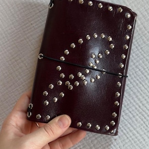 Peut inclure: Une couverture de carnet en cuir bordeaux foncé ornée de clous métalliques argentés disposés le long des bords et suivant une courbe sur le devant. Un fin élastique noir entoure horizontalement le centre pour maintenir la couverture fermée.