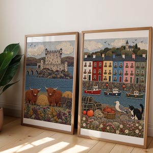 May include: Two framed art prints featuring detailed illustrations. The left print shows a stone castle on an island, two highland cows, and wildflowers. The right print depicts a colorful row of houses, fishing boats, a seagull, and a cat.