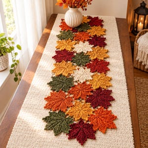 May include: A rectangular cream-colored table runner featuring a central decorative arrangement of crocheted maple leaves in shades of dark green, mustard yellow, burnt orange, and deep burgundy, creating a textured autumn-inspired pattern.