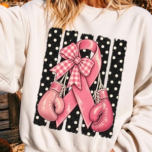 Op de afbeelding: Een crèmekleurig sweatshirt met lange mouwen en een grafische print van een roze bewustwordingslint met een roze-witte gingham strik, geflankeerd door twee roze bokshandschoenen tegen een achtergrond van zwarte verticale strepen met witte stippen.