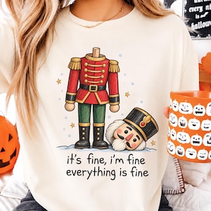 May include: A cream-colored crewneck t-shirt featuring a graphic of a headless nutcracker standing next to its own head on the ground. The text below reads, "it's fine, i'm fine everything is fine." The design includes small gold stars and blue dots.