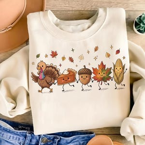 Peut inclure: Un sweat-shirt à col rond de couleur crème avec une impression graphique représentant une dinde, une part de tarte à la citrouille, un gland, une feuille d'érable et un épi de maïs se tenant la main. Le motif inclut des feuilles d'automne.