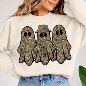 Peut inclure: Un sweat-shirt à col rond couleur crème arborant un motif de trois fantômes avec un imprimé camouflage marron et beige. Le fantôme central porte un bob camouflage et un collier avec plusieurs petits sifflets de chasse marron.