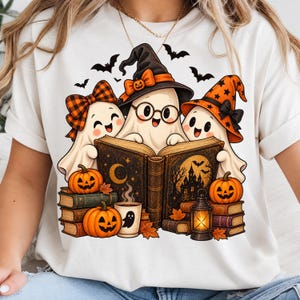Peut inclure: Un t-shirt blanc avec une illustration de trois fantômes de dessin animé lisant un grand livre. Les fantômes portent des chapeaux et des nœuds, entourés de citrouilles, de piles de livres, d'une lanterne et de chauves-souris aux tons orange.