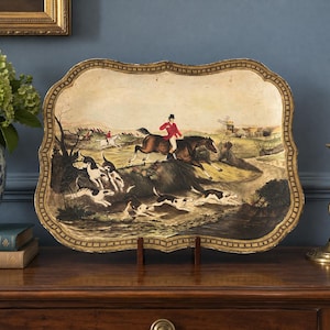 May include: A rectangular decorative tray with a scalloped edge and a gold-toned frame. The center features a painted scene of a rider in a red coat on a brown horse jumping over a hedge, accompanied by a pack of hounds in a landscape.