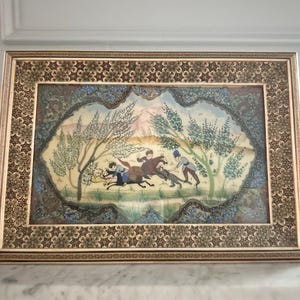 May include: A rectangular framed painting featuring a miniature scene of figures on horseback and on foot in a landscape with trees. The frame has a detailed geometric star pattern border in shades of brown, gold, and black.
