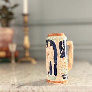 May include: A ceramic beer stein featuring a raised relief design of a figure standing among trees. The vessel is decorated with a color palette of deep blue, cream, and orange accents, and includes a textured handle on the right side.