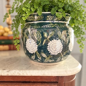 May include: A round ceramic planter with a deep teal glaze featuring hand-painted white chrysanthemum flowers and green foliage patterns. The pot has a wide, flared rim and contains a lush, vibrant green maidenhair fern plant.