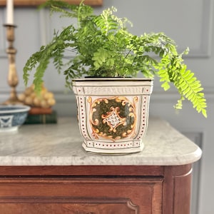 May include: A square ceramic planter featuring a white base with ornate decorative panels in shades of olive green, gold, and brown. The planter has a flared rim and a tapered base, containing a vibrant green fern plant with delicate fronds.