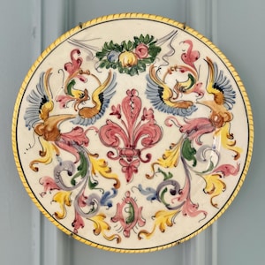 May include: A round ceramic decorative plate featuring a central pink fleur-de-lis motif surrounded by symmetrical hand-painted designs of stylized birds, scrolling acanthus leaves in yellow, blue, and green, and a small fruit cluster at the top.