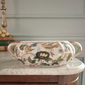 May include: A cream-colored ceramic bowl with two handles, featuring a crackled glaze finish and a decorative pattern of birds, green leaves, and a dark nest containing three yellow eggs. The bowl has a rounded shape with subtle vertical ridges.