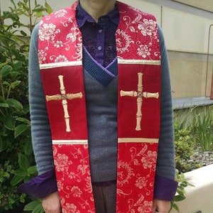May include: A red liturgical stole with a floral pattern and gold accents. The stole features a gold cross design and a gold fringe at the bottom. The stole is worn over a gray sweater and purple shirt.