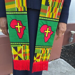 Op de afbeelding: Een groene en bedrukte stola met een rood Afrikaans continentontwerp en een geel kruis. De stola heeft rode franjes aan de onderkant. Het wordt gedragen over een marineblauw kledingstuk.