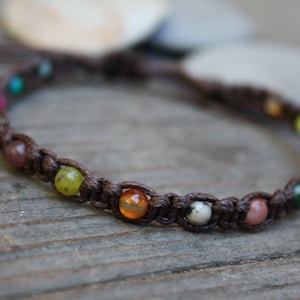 May include: A brown cord bracelet with colorful gemstone beads. The bracelet is made with a knotted cord and features a variety of small, round beads in shades of green, red, orange, yellow, and blue.