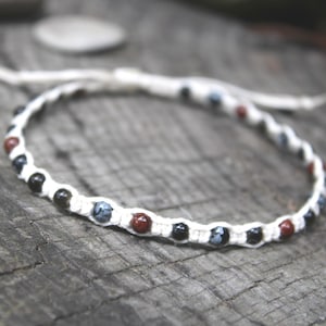 May include: Handmade macrame bracelet featuring a white cord and a series of small, colorful beads. The beads are a mix of black, red, and blue, creating a unique pattern. The bracelet is displayed on a rustic wooden surface.