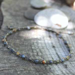 May include: A gray and orange beaded necklace made with macrame cord. The necklace is on a wooden surface.