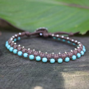 May include: A handmade bracelet featuring a brown woven band adorned with small, light blue beads and silver-colored accents. The bracelet is designed with a clasp closure and is displayed on a wooden surface.