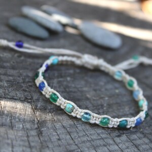 May include: A macrame bracelet crafted from off-white cord featuring a series of small, round beads in various shades of blue, green, and turquoise. The bracelet includes adjustable cord ends and a sliding knot closure for a secure and flexible fit.